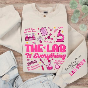 May include: A beige sweatshirt with a pink graphic design that says "The Lab is Everything" and includes various science-related icons. The text "Get It Spun, Run and Done" is also included in the design.