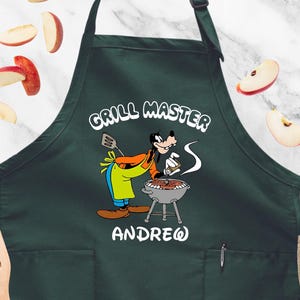 May include: Dark green apron with the words "GRILL MASTER" and a cartoon character grilling. The apron has a pocket and the name "ANDREW" below the character. Slices of apples are scattered around the apron.