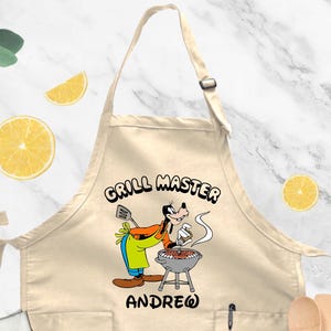 May include: A beige apron with a cartoon image of a dog grilling. The text "GRILL MASTER" is above the dog, and the name "ANDREW" is below. The apron has two pockets and a pen holder.
