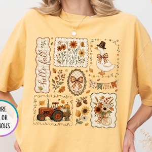 May include: A mustard yellow t-shirt featuring a graphic print with the text "hello fall". The design includes a tractor, a goose wearing a hat, floral arrangements, a bow, and autumn leaves arranged in rectangular and oval frames.