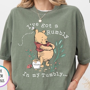 May include: A moss green t-shirt featuring a graphic of Winnie the Pooh with a honey pot, a ladybug, and green foliage. The text reads, I've got a Rumbly in my Tumbly. The shirt has short sleeves and a crew neckline, with a small bee flying above.