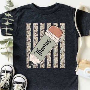 May include: A dark grey heather t-shirt featuring a graphic of a pencil with the name Thomas written on it, positioned diagonally over a vertical camouflage striped background. The shirt is short-sleeved with a crew neckline and a soft fabric texture.