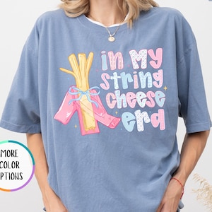 May include: A blue jean colored t-shirt with the phrase "In My String Cheese Era" in pink, blue, and yellow lettering. The graphic includes a bundle of string cheese with pink ribbons. The shirt has short sleeves and a relaxed fit.
