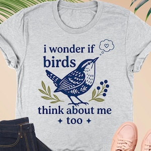 May include: An athletic heather gray short-sleeve t-shirt featuring a dark blue graphic of a bird with the text "i wonder if birds think about me too" and a small thought bubble with a heart, accompanied by dark blue denim jeans and pink sneakers.
