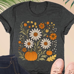 May include: Dark grey heather t-shirt with a floral and pumpkin design. The shirt is paired with dark wash jeans, a pink wallet, and pink sneakers. The shirt's design includes white daisies, orange flowers, and pumpkins.