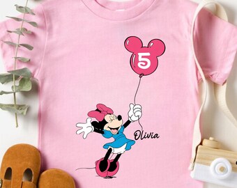 Könnte beinhalten: Rosafarbenes T-Shirt mit Minnie Mouse, die einen Ballon mit der Zahl 5 hält. Das T-Shirt hat kurze Ärmel und einen Rundhalsausschnitt. Der Name "Olivia" ist darunter aufgedruckt. Braune Babyschuhe aus Leder liegen daneben.