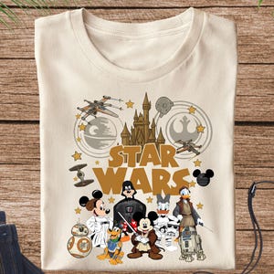 May include: Cream-colored t-shirt featuring a Star Wars graphic with Disney characters. The design includes the Star Wars logo, a castle, spaceships, and characters like Mickey Mouse, Darth Vader, and Donald Duck. The shirt is paired with blue jeans.