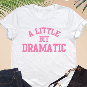 a t - shirt that says a little bit dramatic next to a pair of glasses