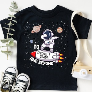 May include: Black t-shirt with a graphic of an astronaut riding a rocket with the text "To Second Grade and Beyond".