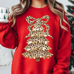 May include: A red sweatshirt featuring a Christmas tree design with a leopard print pattern. The tree is adorned with a bow at the top. A white mug with green Christmas trees is held in the hand.