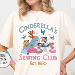 May include: A light beige t-shirt with a graphic of Cinderella's mice sewing. The text reads "Cinderella's Sewing Club Est. 1950".