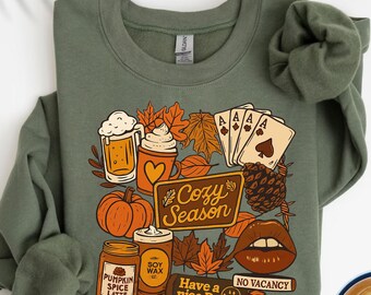 May include: A military green crewneck sweatshirt featuring a graphic print with autumn-themed illustrations including a pumpkin, a beer mug, a coffee cup, playing cards, a pinecone, and a pair of lips, with text reading Cozy Season.