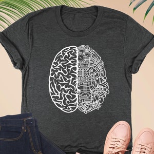 May include: A dark heather grey t-shirt with a white graphic of a brain with gears and cogs on one side and a human brain on the other side. The shirt is shown on a model with blue jeans and pink sneakers.