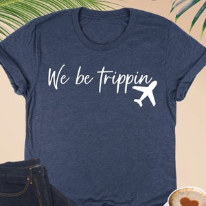 K&ouml;nnte beinhalten: Ein heidefarbenes marineblaues T-Shirt mit wei&szlig;em Text, der "We be trippin'" lautet, und einer wei&szlig;en Flugzeuggrafik.