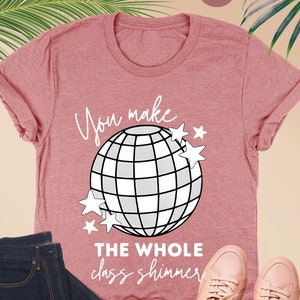 May include: Heather mauve t-shirt with a disco ball graphic and the text "You make the whole class shimmer." The shirt is paired with dark wash jeans, clear-framed glasses, and pink sneakers.