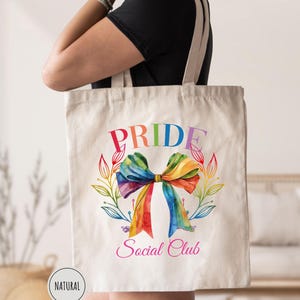May include: A natural canvas tote bag featuring the word "PRIDE" in rainbow colors above a watercolor bow. The bow is in rainbow colors, with the words "Social Club" below. The bag has long, natural-colored straps.