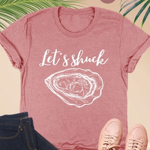 May include: A heather mauve t-shirt with the text "Let's Shuck" and a white outline drawing of an oyster.