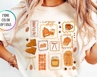 May include: Ivory-colored t-shirt with a fall-themed graphic. The design includes illustrations of boots, a pumpkin, a coffee cup, a hat, and the word "autumn". The shirt is made of a soft material.