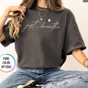May include: A gray t-shirt with the text "just breathe" in white script with a small white heart above the text. The model is wearing the shirt with blue jeans and black sneakers.