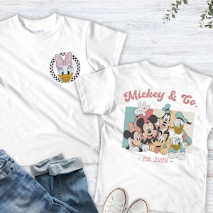 May include: Two white t-shirts with Disney character designs. One shirt features Daisy Duck, the other has Mickey & Co. with Mickey Mouse, Minnie Mouse, Donald Duck, and Goofy. Blue jeans and white sneakers are also in the image.
