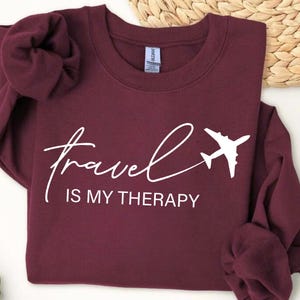 Pu&ograve; includere: Felpa bordeaux con la scritta bianca "travel is my therapy" e una grafica di aereo bianca. Il top girocollo a maniche lunghe &egrave; esposto su una superficie bianca.