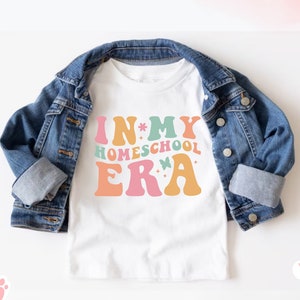 May include: White t-shirt with the text "IN MY HOMESCHOOL ERA" in colorful, retro-style lettering. The shirt is paired with a denim jacket. The design includes butterflies and floral accents, creating a cheerful aesthetic.