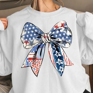 May include: White sweatshirt featuring a large bow design with patriotic patterns. The bow incorporates blue with white stars, red and white stripes, and a leopard print. The sweatshirt is a casual style, suitable for everyday wear.