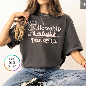 May include: A gray t-shirt with a vintage-style graphic that says "Fellowship Delivery Co." with the text "Est. 1954" and "fast, reliable, precious" underneath. The graphic features a line of small figures walking through a forest.
