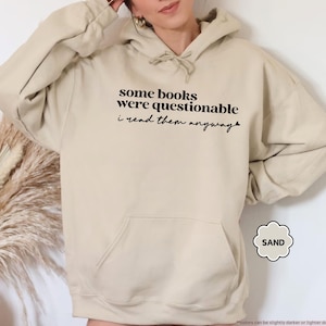 May include: A sand-colored hooded sweatshirt featuring a front kangaroo pocket and a drawstring hood. The chest displays black text that reads: some books were questionable i read them anyway, with a small heart icon at the end of the sentence.