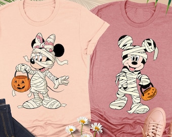 May include: Two short-sleeve crew neck t-shirts in heather peach and heather mauve. The peach shirt features a graphic of a mummy Minnie Mouse holding a jack-o'-lantern. The mauve shirt features a mummy Mickey Mouse holding a jack-o'-lantern.