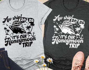 May include: Two t-shirts, one light gray and one dark gray, with a cruise ship graphic and the text "Aw ship! It's our Honeymoon Trip 2026". The shirts are displayed with a pair of blue jeans and pink sneakers.