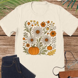 May include: Cream-coloured t-shirt with a floral design featuring daisies, orange flowers, and pumpkins. The design is centered on the shirt. The shirt is paired with dark blue jeans and a brown purse with glasses.