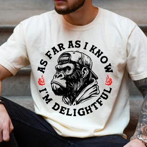 May include: Ivory-colored t-shirt featuring a black and white graphic of a gorilla wearing a cap. The text around the gorilla reads "AS FAR AS I KNOW I'M DELIGHTFUL" in a circular design. The shirt is a casual wear item.