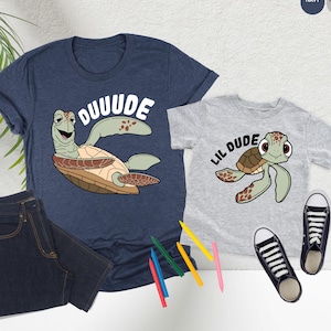 May include: Two matching graphic t-shirts, one navy and one heather gray, featuring cartoon sea turtles. The navy shirt says "DUUUDE" and the gray shirt says "LIL DUDE". Dark blue jeans and black sneakers complete the casual ensemble.