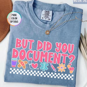 May include: Blue jean colored t-shirt with the text "BUT DID YOU DOCUMENT?" in pink and white. The shirt has medical-themed graphics, including a coffee cup, bandage, stethoscope, heart, star of life, syringe, flower, and a pill. A gold necklace is draped across the shirt.