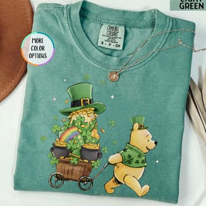 May include: Light green t-shirt featuring a St. Patrick's Day graphic. The design includes a leprechaun with a pot of gold and a bear pulling a cart. The shirt has a crew neck and short sleeves. The text "LIGHT GREEN" is in the upper right corner.