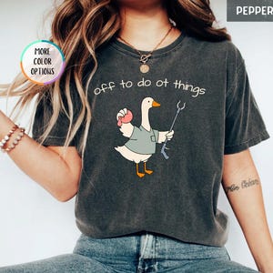 May include: A charcoal gray t-shirt featuring a graphic of a white goose wearing a gray shirt, holding a pink object and a reacher grabber tool. The text above the goose reads, off to do ot things. The shirt has a short-sleeved, crew-neck design.