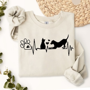 May include: A beige sweatshirt with a black paw print, heartbeat, cat, and dog silhouette design. The design includes three small hearts. The text "SAND" is visible in the upper right corner of the image.