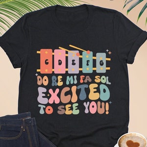 May include: Black t-shirt with a colorful xylophone graphic and the text "DO RE MI FA SOL EXCITED TO SEE YOU!"