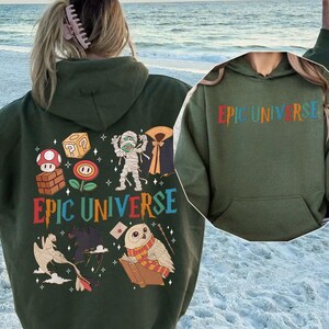 May include: A military green hoodie with the text "EPIC UNIVERSE" in colorful letters. The back of the hoodie features various cartoon graphics, including a mushroom, a cube, a mummy, and an owl reading a book.