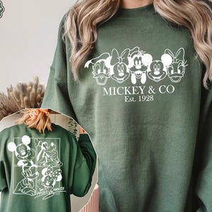 May include: Military green sweatshirt featuring a white graphic of Mickey Mouse and friends. The front design includes the text "MICKEY & CO Est. 1928". The back of the sweatshirt also has a white graphic of Mickey Mouse and friends.