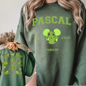 Op de afbeelding: Militairgroen sweatshirt met de naam "PASCAL" in lichtgroen, een cartoon kikkerfiguur en de tekst "TANGLED" en "2010". De achterkant van de trui toont meerdere kikker illustraties.