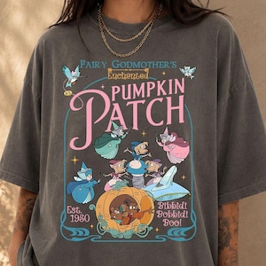 May include: A charcoal gray short-sleeve t-shirt featuring a graphic print with the text Fairy Godmother's Enchanted PUMPKIN PATCH, Est. 1950, and Bibbidi Bobbidi Boo. The design includes illustrations of mice, a pumpkin carriage, and a glass slipper.