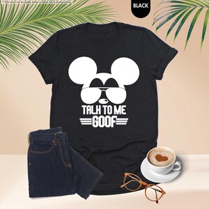 May include: Black t-shirt featuring a white Mickey Mouse graphic wearing sunglasses and the text "TALK TO ME GOOF". The image includes dark wash jeans, a coffee cup, and a pair of spectacles.