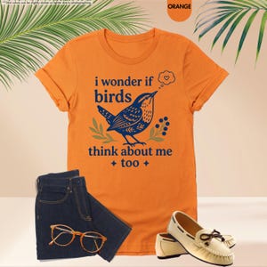 May include: An orange short-sleeved t-shirt featuring a dark blue graphic of a bird, green leaves, and the text "i wonder if birds think about me too" with a small heart in a thought bubble, paired with dark blue denim shorts, glasses, and loafers.