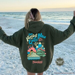 Pu&ograve; includere: Una felpa con cappuccio verde con una stampa grafica del castello di Disneyland e il testo "The Happiest Place on Earth".