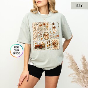 May include: A sage green short-sleeve t-shirt featuring a graphic print with the text hello fall. The design includes a goose wearing a hat, a red tractor, autumn flowers, pumpkins, a plaid bow, and bunting flags arranged in a collage style.