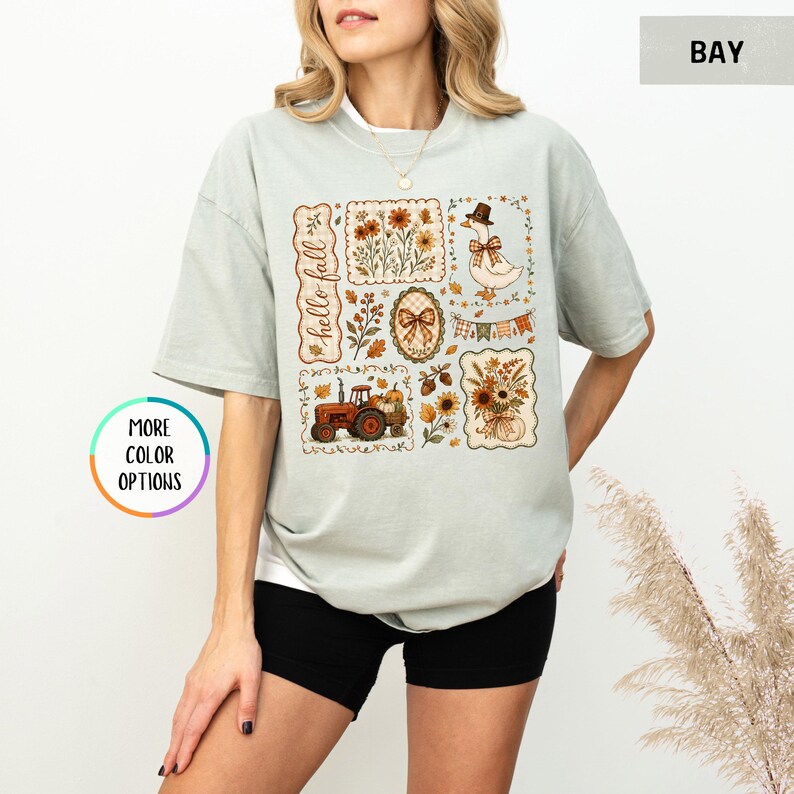 May include: A sage green short-sleeve t-shirt featuring a graphic print with the text hello fall. The design includes a goose wearing a hat, a red tractor, autumn flowers, pumpkins, a plaid bow, and bunting flags arranged in a collage style.