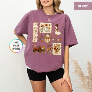 May include: A berry-colored short-sleeve t-shirt featuring a graphic print with the text "hello fall" alongside illustrations of a tractor, a goose wearing a hat, sunflowers, autumn leaves, a plaid bow, and a string of pennant flags.