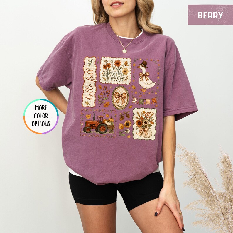 May include: A berry-colored short-sleeve t-shirt featuring a graphic print with the text "hello fall" alongside illustrations of a tractor, a goose wearing a hat, sunflowers, autumn leaves, a plaid bow, and a string of pennant flags.